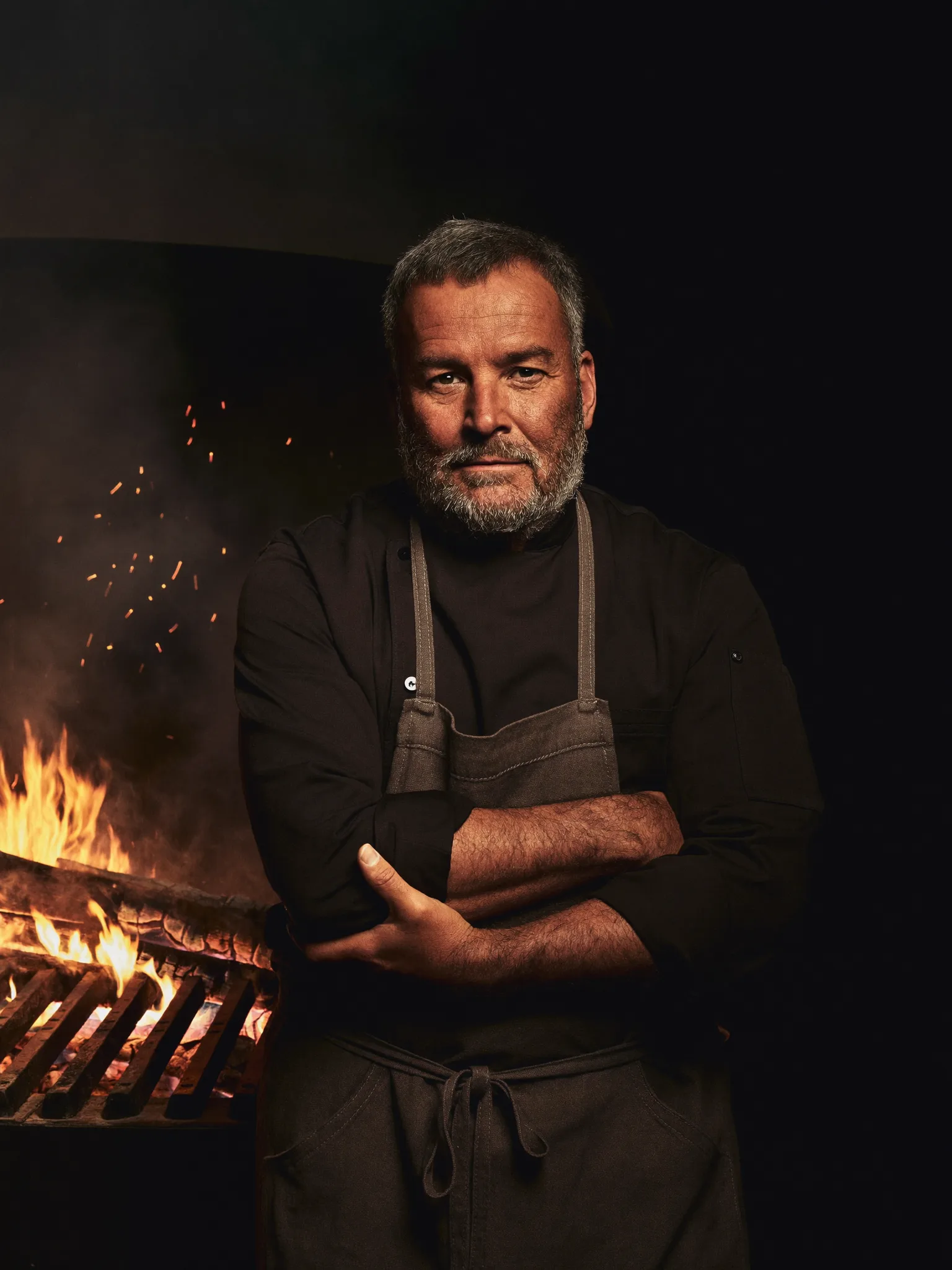 Chef Ilmar Kask in near-darkness beside the hearth, his face lit by fire glow, holding a long iron poker.